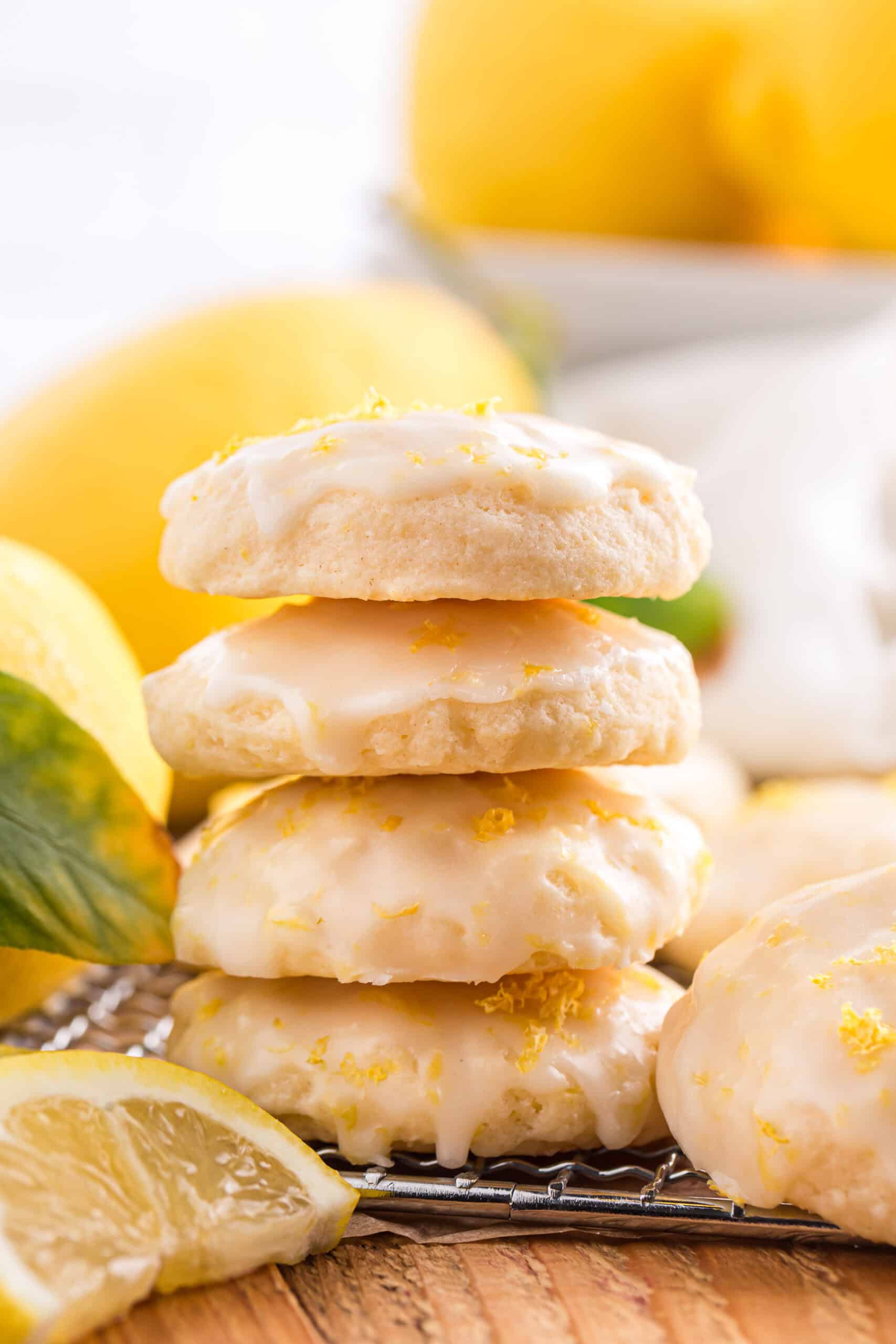 Stack of lemon ricotta cookies with lemon glaze and zest, surrounded by fresh lemons on a wooden board