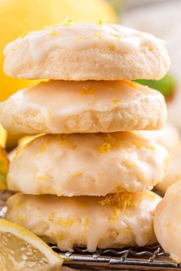 Stack of lemon ricotta cookies with lemon glaze and zest, surrounded by fresh lemons on a wooden board