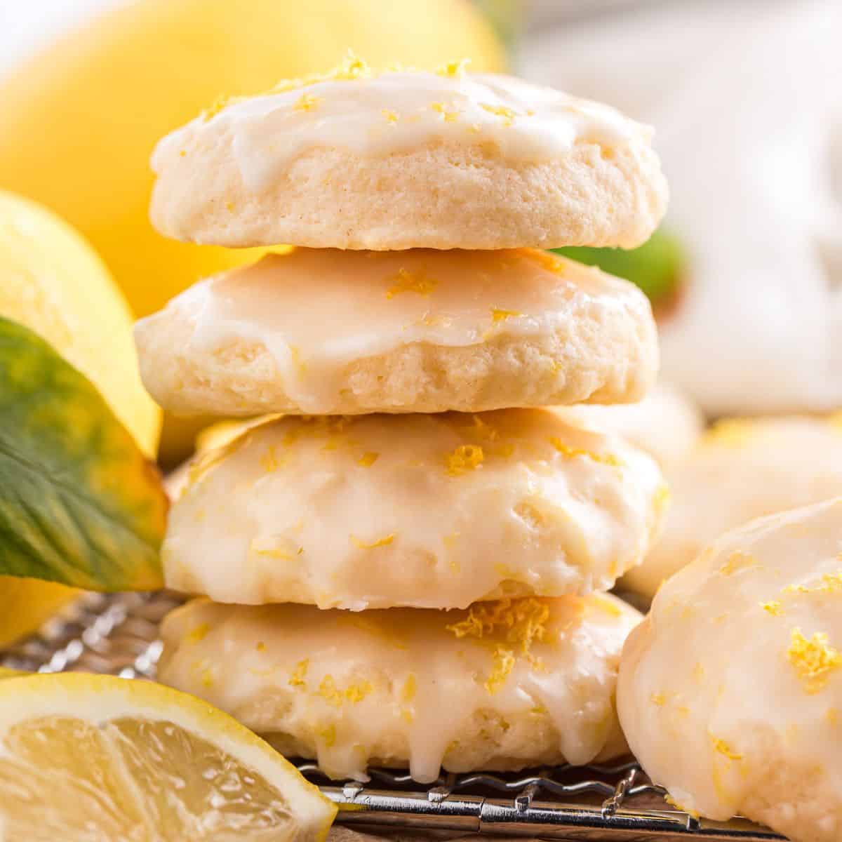 Stack of lemon ricotta cookies with lemon glaze and zest, surrounded by fresh lemons on a wooden board
