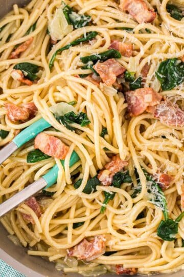 Close-up of bacon and spinach pasta being mixed in the skillet with tongs.